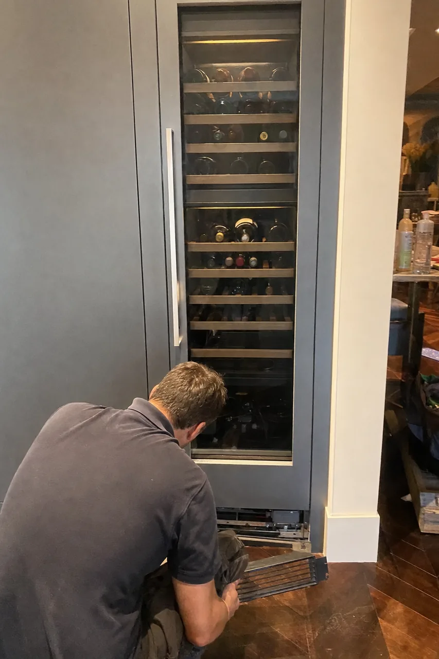 A full-height integrated wine column built into cabinetry, bottles visible under low interior light, a technician crouched at its base holding the lower grille.