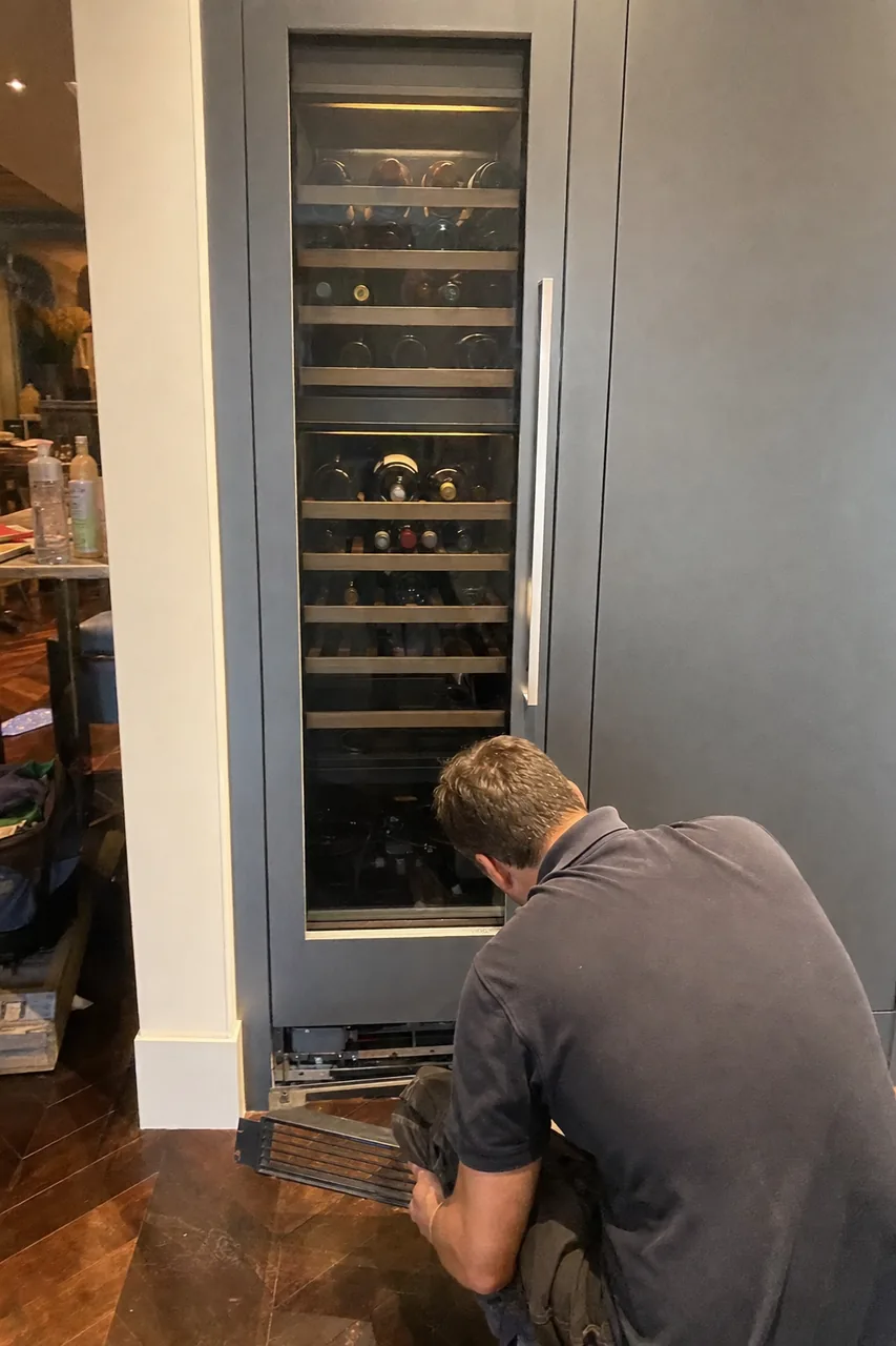 Mirrored view of the integrated column, its glass door closed on the racked bottles and the removed grille resting against the plinth.