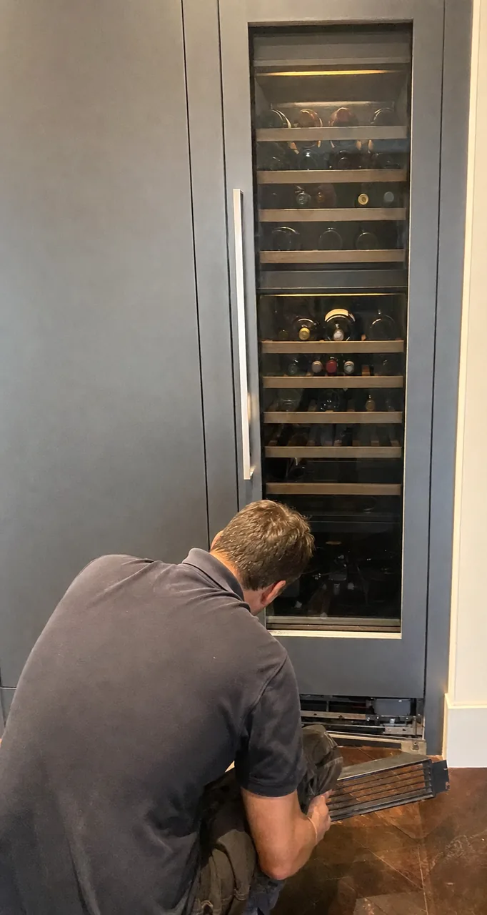 Close crop of the wine column, bottles racked in low light behind the glass and the technician’s shoulder at the frame edge.