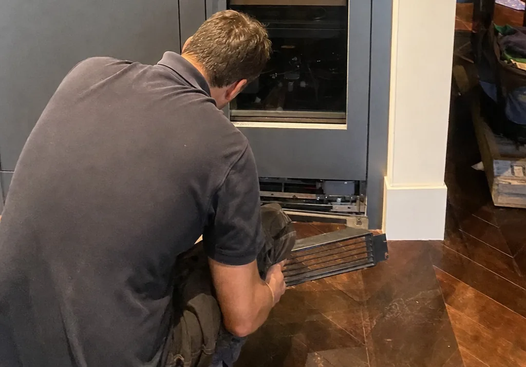 Wide crop at the foot of the built-in column: a kneeling technician lifting the base grille clear of the machine compartment, the cabinet plinth and wood floor beside him.