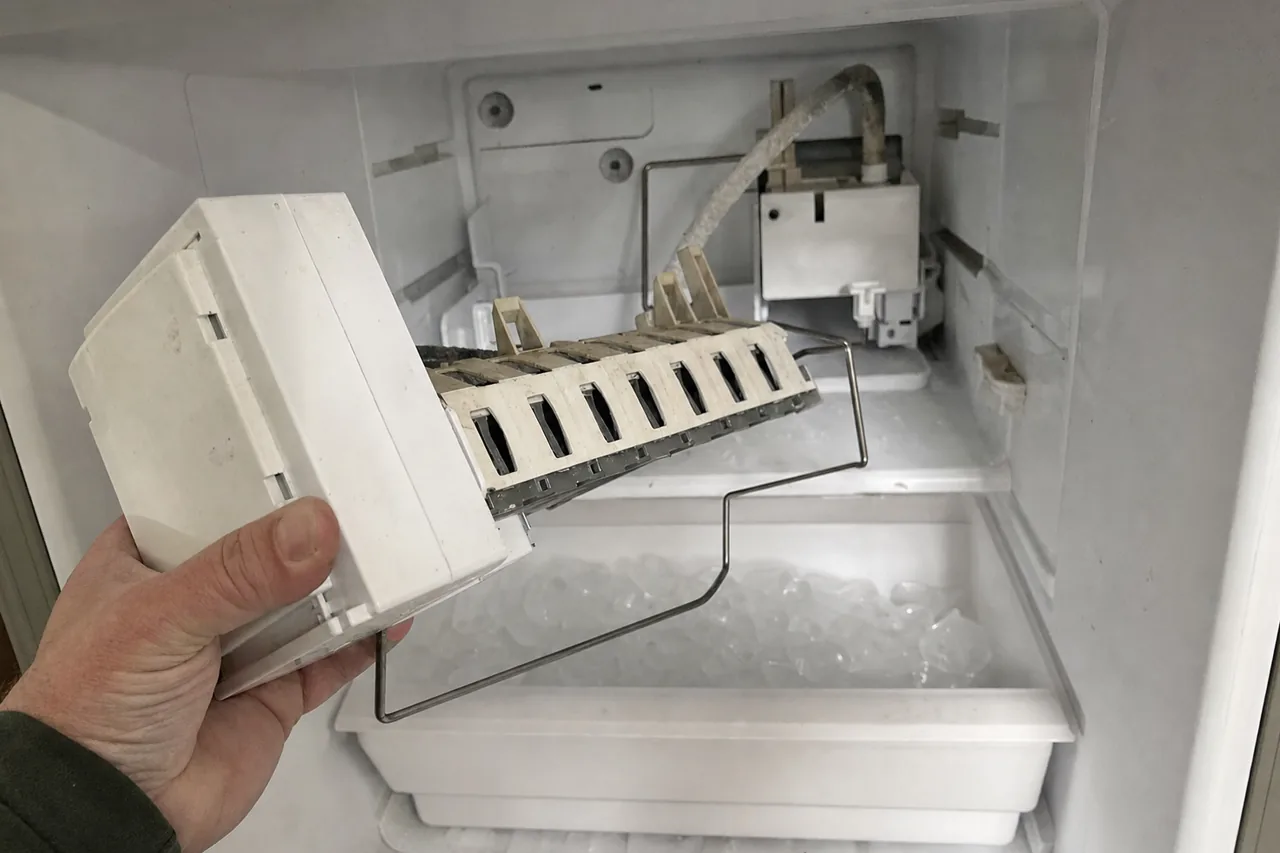 An ice maker module held clear of the freezer compartment in one hand, its harvest arm and white housing forward of the empty mounting bracket.