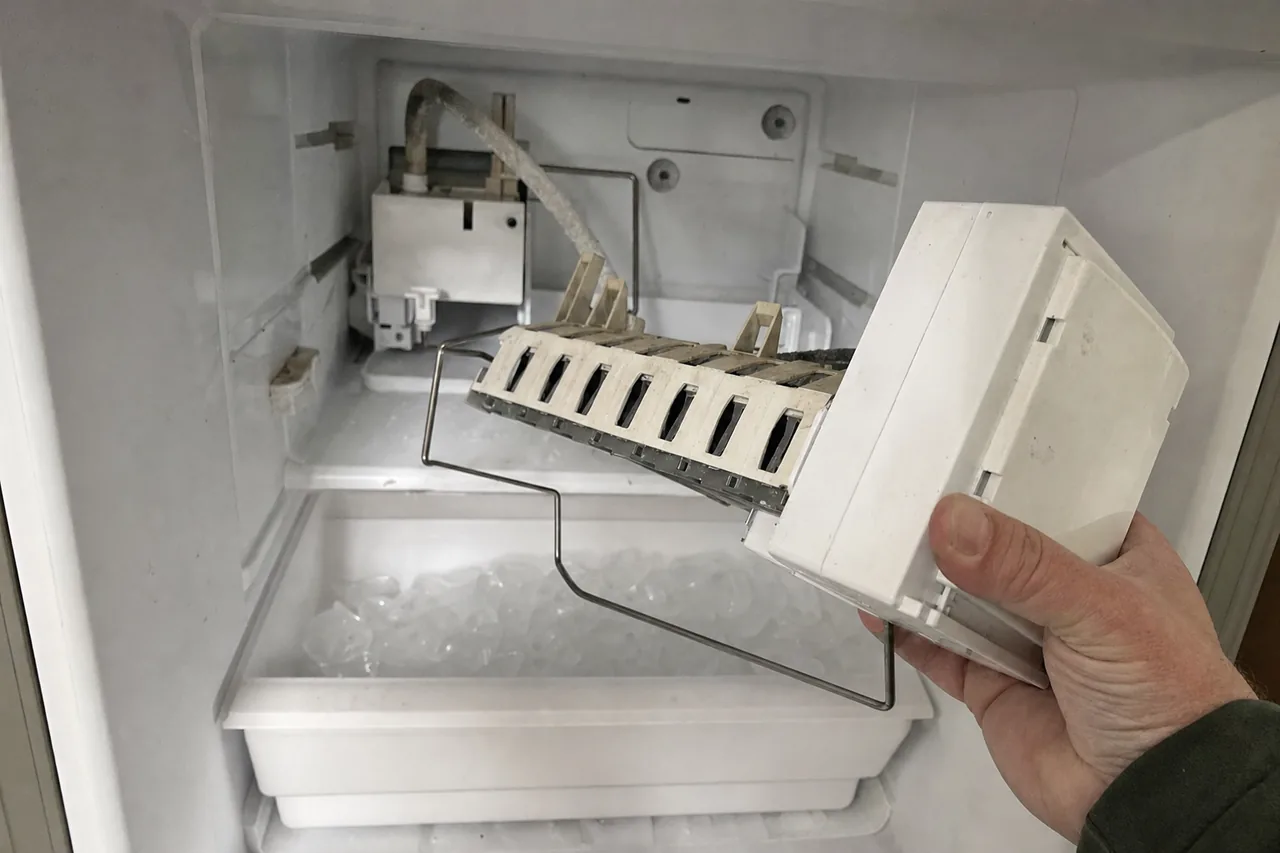 Mirrored view of the removed ice maker module, the water fill tube behind it and melting ice in the bin below.