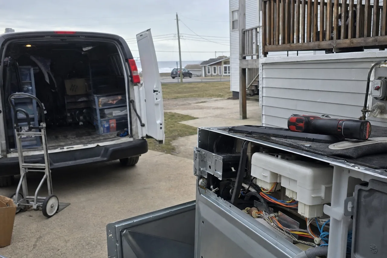 A plain white service van parked in a residential driveway under grey sky, rear doors open on stacked plastic parts bins and a folded hand truck.
